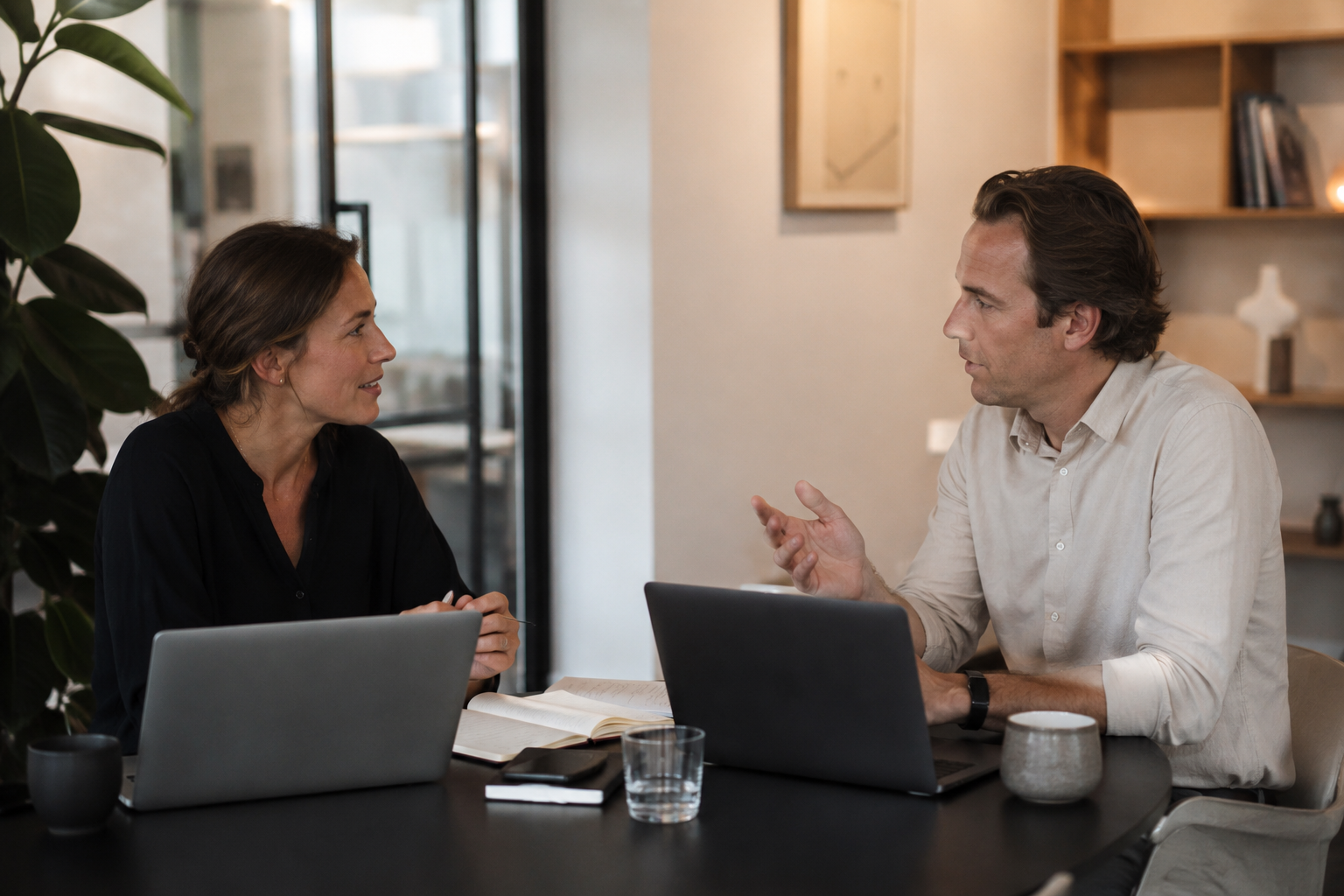 Twee professionals voeren een ontspannen gesprek aan een vergadertafel met laptops en notitieboek